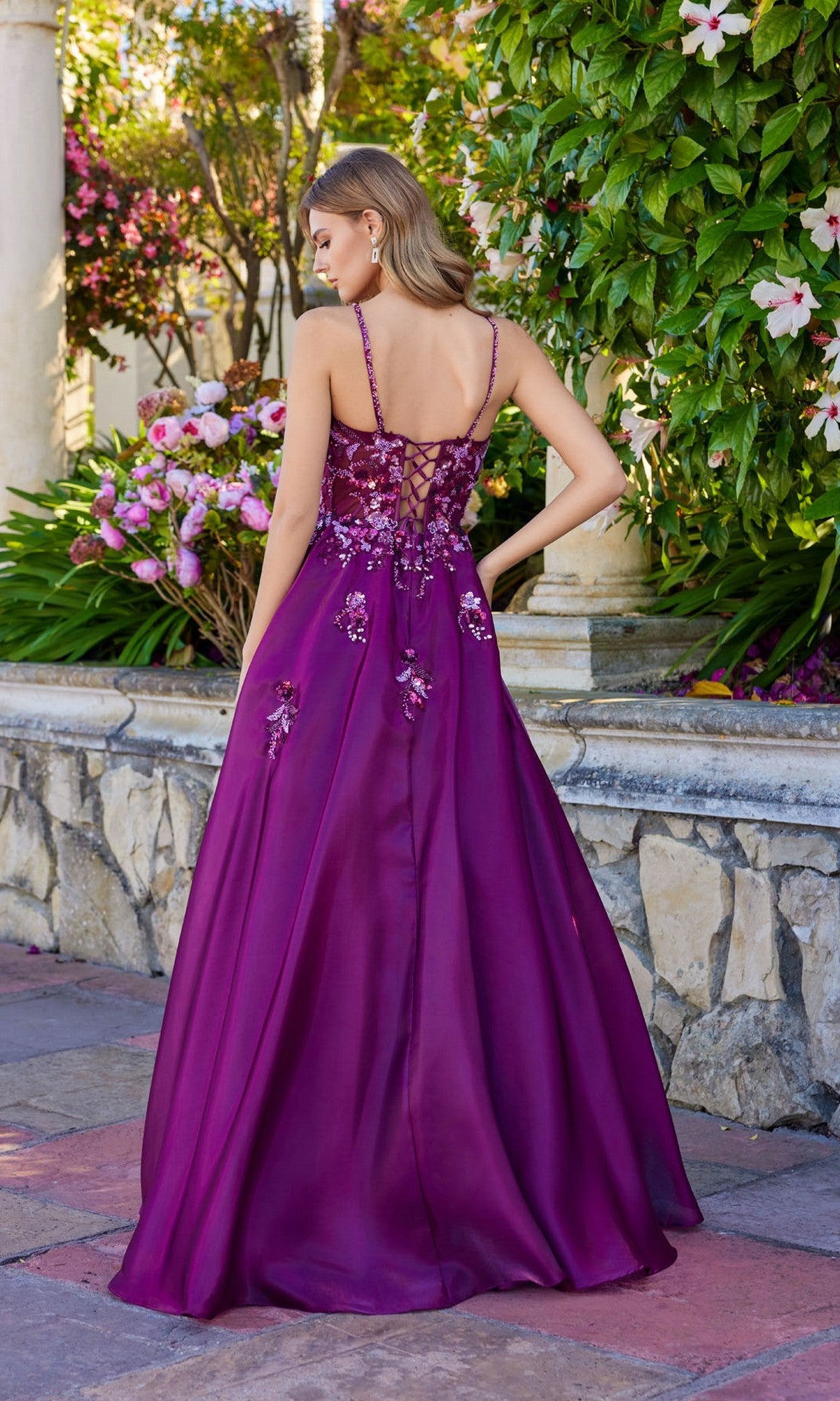 A woman stands outdoors near stonework and flowering plants, facing away, wearing the Juliet JT2617U—a sleeveless, floor-length purple A-line prom dress with floral embroidery and a lace-up back.